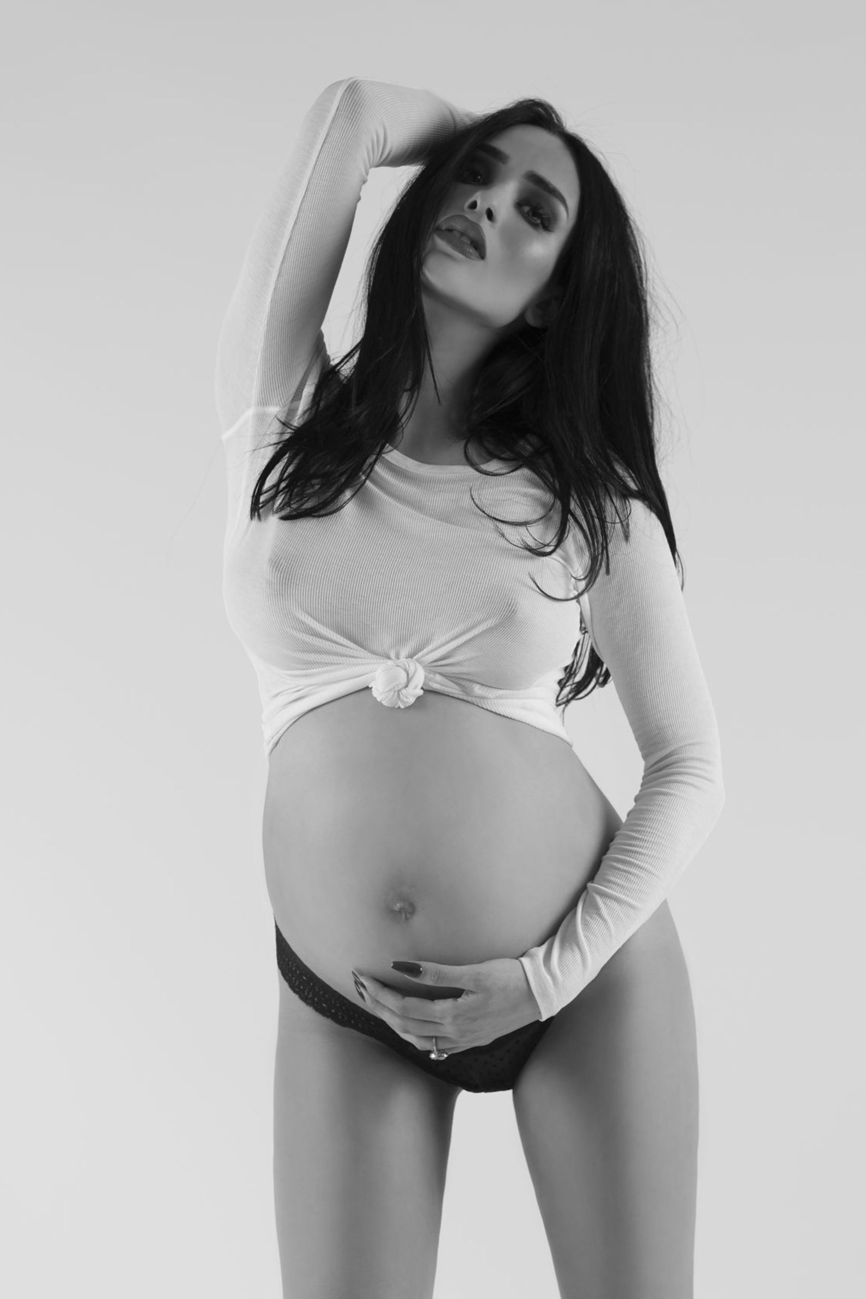 Black and white maternity photo. A woman posing sensually with her left hand on her exposed stomach and her right on the back of her head, which is tilted back.