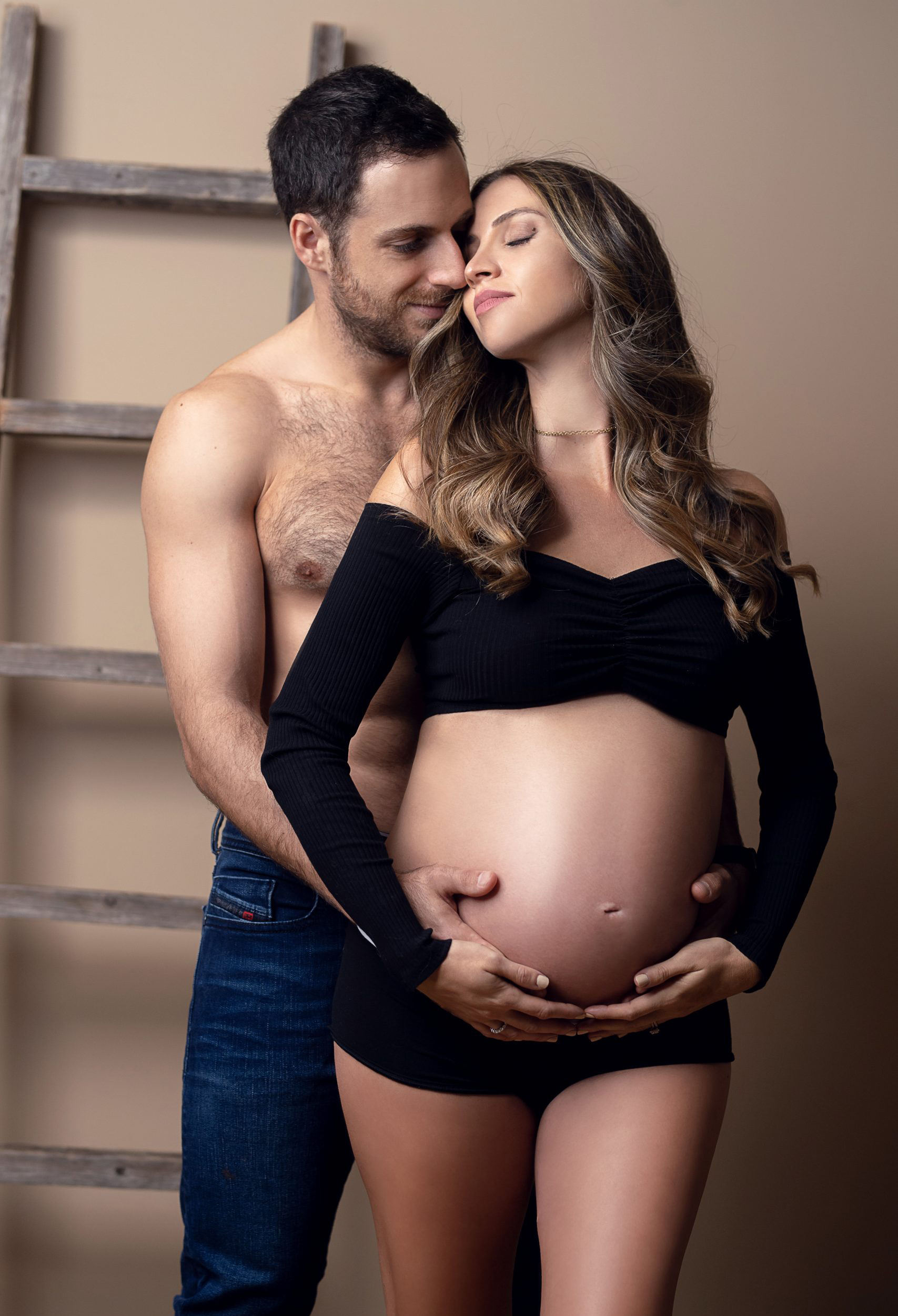 A couple in a maternity picture. The woman's belly is exposed, and the man is shirtless with his hand resting on her belly.